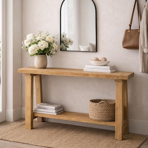 May include: A wooden console table with a lower shelf, topped with books, a woven basket, and a vase of white flowers. A black-framed arched mirror hangs above the table. A woven rug sits beneath the table.