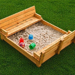 May include: A wooden sandbox with a built-in bench, filled with sand. Colorful plastic toys, including a red ball, a blue cup, and a green cup, are inside the sandbox. The sandbox is set on a green lawn.