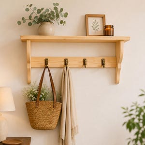 May include: A light-colored wooden wall shelf with four hooks, holding a woven basket with flowers and a beige scarf. The shelf also displays a framed picture, a candle, and a vase with greenery. The shelf is mounted on a white wall.