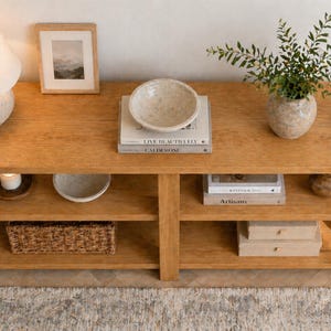Könnte beinhalten: Eine hellbraune Holzkonsolentisch mit zwei Regalen, auf der dekorative Gegenstände ausgestellt sind. Oben befinden sich eine Lampe, ein gerahmtes Bild, eine Schale, Bücher und eine Vase mit Grünzeug. Ein geflochtener Korb und eine Kerze befinden sich auf den Regalen.
