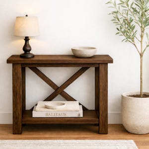 Może przedstawiać: Ciemnobrązowa drewniana konsola z podporą w kształcie litery X. Na górze lampa z beżowym kloszem i dekoracyjna miska. Na dolnej półce stos książek z napisem "BALITAUS". Po prawej stronie roślina doniczkowa.