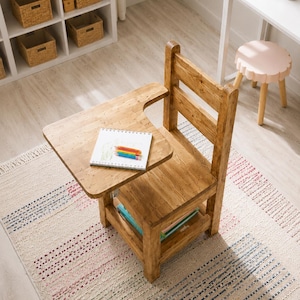 Op de afbeelding: Houten schoolbureau stoel met een vastgeschroefd schrijfoppervlak. Een notitieboek en kleurpotloden liggen op het bureau. De stoel staat op een patroonkleed met blauwe, rode en roze strepen. Een rek en een kleine roze kruk staan op de achtergrond.
