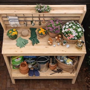 Puede incluir: Un banco de trasplante de madera con varios suministros de jardinería. El banco contiene flores en macetas, guantes de jardinería, herramientas y una regadera. El banco es de madera clara y está al aire libre.