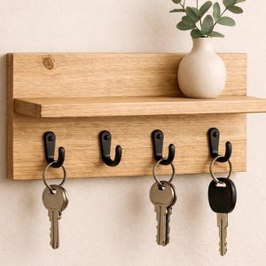 May include: Wooden key holder with a small shelf and four black hooks. The key holder has a light brown finish and holds several keys. A small, off-white vase with green foliage sits on the shelf.