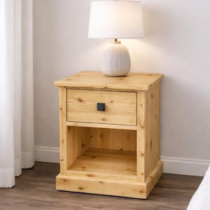 May include: A light-coloured wooden bedside table with a drawer and open shelf. A white lamp with a textured base and a white lampshade sits on top. The nightstand has a black square drawer pull.