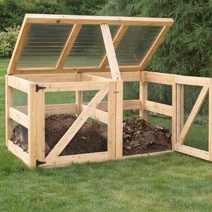 Pode incluir: Um recipiente de compostagem de madeira com uma tampa articulada com painéis transparentes e uma porta parcialmente aberta. O recipiente é construído com madeira de cor clara e apresenta laterais de malha de arame. Está sobre relva verde.