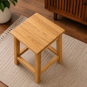 May include: A square wooden stool with a natural finish. The stool has four legs and a flat top. It is sitting on a beige rug with fringe. A potted plant is in the background.