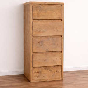 Könnte beinhalten: Eine hohe Holzkommode mit fünf Schubladen. Das helle Holz weist sichtbare Maserungen und Knoten auf. Sie steht vor einer weißen Wand und ruht auf einem Holzboden.