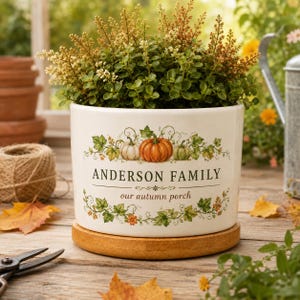 Op de afbeelding: Een witte keramische plantenpot met een houten voetstuk en een groene plant. De pot is bedrukt met een illustratie van drie pompoenen tussen groene ranken en de tekst "ANDERSON FAMILY" boven "our autumn porch" in het zwart.