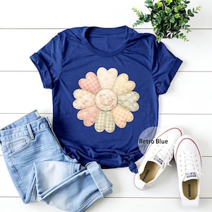 以下が含まれることがあります： 大きな花柄が特徴のネイビーブルーのTシャツ。花はギンガムチェックや花柄など、さまざまな柄の生地で作られています。明るいブルーのジーンズと白いスニーカーと合わせています。