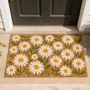 May include: A rectangular doormat with a daisy floral pattern. The mat features white daisies with yellow centers and green stems and leaves against a brown background. The mat is placed in front of a dark door.