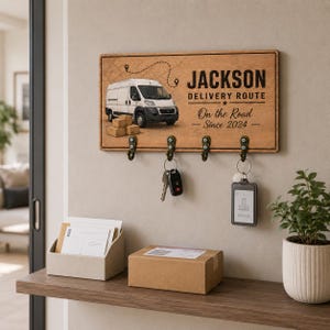 May include: A rectangular wooden wall-mounted key holder featuring a graphic of a white delivery van, a dotted route line, and text reading JACKSON DELIVERY ROUTE On the Road Since 2024. It includes four dark metal hooks with keys attached.