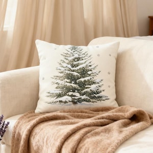 May include: A square decorative pillow with a winter scene. The pillow features a detailed illustration of a snow-covered evergreen tree in shades of green and white. Small snowflake accents are scattered around the tree on the off-white background. The pillow is placed on a beige sofa with a brown blanket.