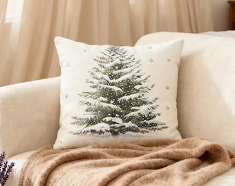 May include: A square decorative pillow with a winter scene. The pillow features a detailed illustration of a snow-covered evergreen tree in shades of green and white. Small snowflake accents are scattered around the tree on the off-white background. The pillow is placed on a beige sofa with a brown blanket.