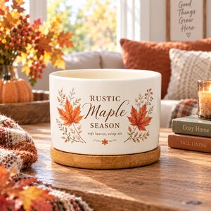 May include: A white ceramic candle vessel with a wooden base, featuring a printed design of orange maple leaves and the text "RUSTIC Maple SEASON soft leaves, crisp air". The cylindrical container has a smooth finish and autumn-themed botanical illustrations.