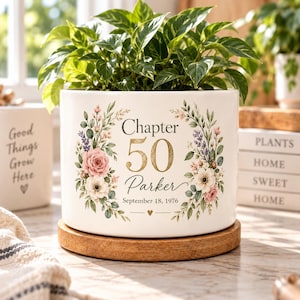 May include: A white ceramic cylindrical planter featuring a floral wreath design with pink and white flowers, green leaves, and gold text that reads Chapter 50 Parker September 18, 1976. The planter sits on a circular wooden base and holds a green plant.