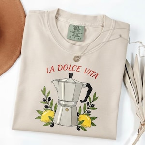 Pode incluir: Camisola bege Comfort Colors com a frase "LA DOLCE VITA" em vermelho. O gráfico apresenta uma cafeteira prateada, limões, azeitonas e ramos de oliveira. Um colar de prata com um pequeno pingente também é visível.