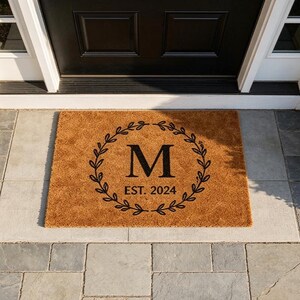 May include: A rectangular brown doormat with a black wreath design around the letter "M" and the text "EST. 2024". The doormat is placed in front of a black door on a gray tiled surface.