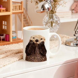 May include: White ceramic coffee mug featuring a detailed illustration of an adorable sea otter. The sea otter has brown fur and a light-colored face. The mug is filled with a warm beverage.