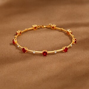 Puede incluir: Una pulsera de cadena dorada que presenta una serie de piedras preciosas rojas redondas alternadas con piedras blancas transparentes más pequeñas dispuestas en círculo. La pulsera incluye un cierre metálico seguro en la parte superior.