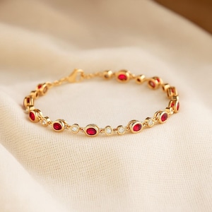 May include: A gold bracelet featuring oval-cut red gemstones and small, round, clear gemstones. The bracelet is set on a soft, off-white fabric. The clasp is visible, and the bracelet is designed with a delicate chain.