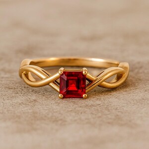 Op de afbeelding: Een goudkleurige ring met een ingeweven ontwerp. De ring heeft een vierkante, helderrode edelsteen in het midden, die contrasteert met de gouden band. Het ontwerp van de ring is elegant en opvallend.