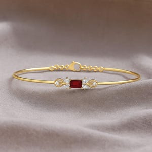 Pode incluir: Uma pulseira rígida de tom dourado com uma pedra vermelha retangular central, ladeada por duas pequenas pedras transparentes de cada lado. A banda metálica é fina e inclui uma pequena corrente de extensão com fecho lagosta na parte traseira.