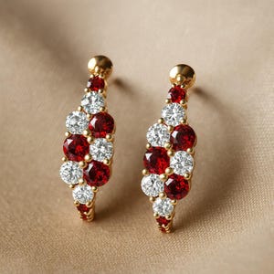 May include: A pair of gold earrings featuring a vertical arrangement of alternating round-cut red and clear white stones. Each earring is designed with a post backing and a slender, elongated shape that showcases the faceted gems in a cluster setting.