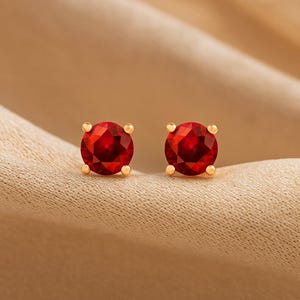 May include: A pair of gold stud earrings, each featuring a round, faceted, deep red gemstone. The earrings are set on a gold post with a small, round backing. The gemstones have a brilliant cut, reflecting light.