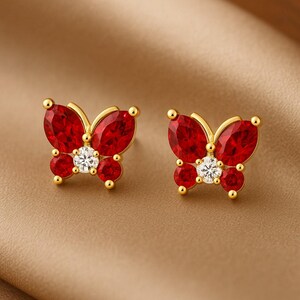 May include: A pair of gold-colored butterfly stud earrings. Each earring features a red, oval-shaped gemstone for the wings, a white, round gemstone for the body, and smaller red gemstones. The earrings have a post backing.