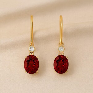 Peut inclure: Une paire de boucles d'oreilles pendantes dorées. Chaque boucle d'oreille présente une grosse pierre précieuse rouge foncé de forme ovale, une petite pierre précieuse ronde et transparente, et un anneau doré. Les boucles d'oreilles sont présentées sur un fond neutre.