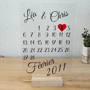 Puede incluir: Un calendario transparente con los nombres "Léa & Chris" en escritura elegante. El calendario presenta un diseño de febrero de 2011, con el día 4 resaltado por un corazón rojo. Se muestra sobre una base de madera.