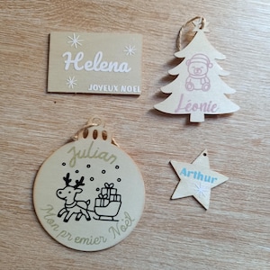 Peut inclure: Ensemble de décorations de Noël en bois. Comprend un ornement rectangulaire avec le nom "Helena" et le texte "JOYEUX NOEL", un ornement en forme d'arbre avec un ours et le nom "Léonie", un ornement rond avec un renne et le nom "Julian", et un ornement en forme d'étoile avec le nom "Arthur".