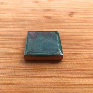 May include: A square, teal decorative tile with a copper-coloured edge. The tile has a glossy finish and is set against a wooden background. The teal colour has darker green and blue hues.