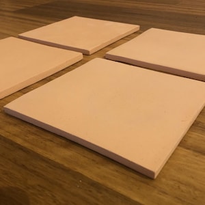 May include: Four square, pale peach ceramic coasters are arranged on a wooden surface. The coasters are evenly spaced, showcasing their simple, minimalist design. The coasters are likely used for protecting surfaces from heat or moisture.