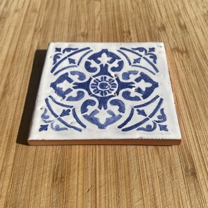 May include: A square ceramic tile featuring a white base with a symmetrical blue floral pattern. The tile has a smooth, glossy finish and a terracotta-coloured edge, displaying a detailed, intricate design centred on the surface of the piece.