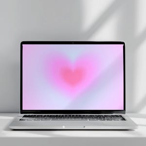 Puede incluir: Un ordenador portátil plateado con teclado negro y una pantalla que muestra una forma de corazón rosa degradado y de enfoque suave sobre un fondo lavanda pálido. El dispositivo cuenta con un marco fino y un acabado metálico, visto de frente.