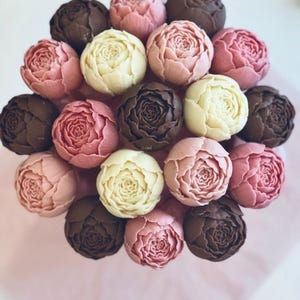 Puede incluir: Un ramo de caramelos de chocolate con forma de flor en tonos rosa, marrón y blanco. Los caramelos están dispuestos en un patrón circular sobre papel rosa, asemejándose a un arreglo floral. Un dulce.
