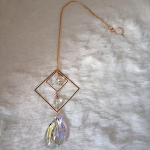 Pode incluir: Um apanhador de sol de metal dourado com uma moldura fina em forma de losango suspensa por uma corrente delicada com um gancho. Duas pequenas contas facetadas transparentes pendem dentro da moldura e um grande prisma de cristal iridescente em gota.