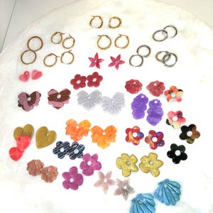 May include: A collection of assorted earrings including gold and silver metal hoops, and various colourful resin shapes such as hearts, flowers, stars, and seashells in shades of pink, purple, orange, blue, gold, and black with glitter finishes.