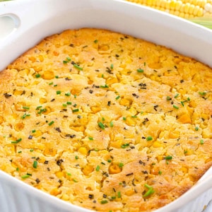 May include: A square, golden-yellow cornbread casserole in a white baking dish, garnished with green chives. Two ears of corn are in the background. The cornbread is speckled with kernels of corn.