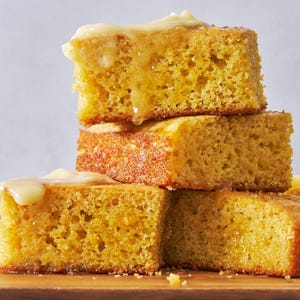 May include: A stack of four golden-yellow cornbread squares, drizzled with a creamy, pale yellow glaze. The cornbread has a porous texture and sits on a wooden surface, creating a visually appealing dessert presentation.