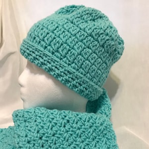 May include: A turquoise crochet hat and scarf set. The hat has a folded brim and a textured pattern. The scarf is draped and also features the same textured crochet design. The set is made of a soft, chunky yarn.