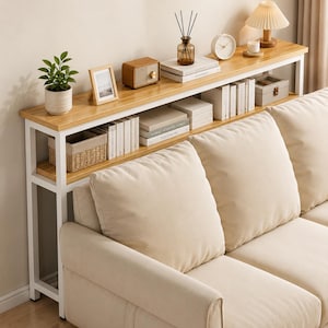 May include: A two-tiered console table with a light wood top and white metal frame. The top shelf displays a small potted plant, books, a radio, a clock, and decorative items. The table is positioned behind a beige sofa.