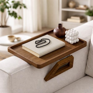 May include: A wooden sofa arm tray with a rectangular design. The tray holds a book, a black beaded necklace, a brown vase, and a white geometric cube. The tray is attached to the sofa arm with a wooden support.