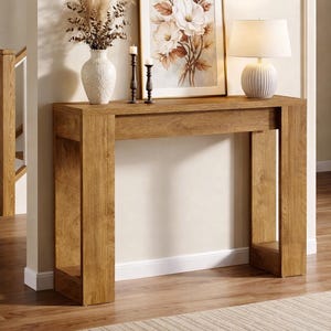 May include: A wooden console table with a framed floral print, a white ceramic vase with dried plants, a white lamp, and two black candle holders. The table is made of light brown wood and sits on a light brown wood floor.