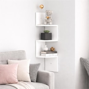 May include: A white corner shelf unit with a zig-zag design. The shelves hold a teddy bear, a potted plant, a vase, books, and decorative objects. A gold-coloured light fixture is mounted above the shelf.