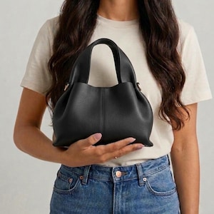 Könnte beinhalten: Eine schwarze Handtasche mit runder Form und zwei kurzen Griffen. Die Tasche wird von einer Person gehalten, die ein helles T-Shirt und eine blaue Jeans trägt. Die Tasche hat eine vertikale Naht in der Mitte.