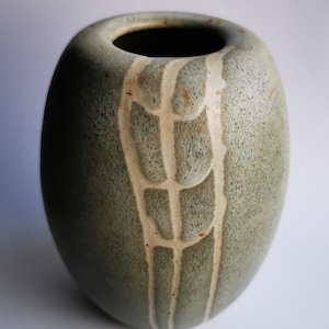 Puede incluir: Un jarrón de cerámica ovalado y moteado en tonos verdes y grises. El jarrón presenta un diseño vertical texturizado en blanco roto. La abertura es oscura, contrastando con el exterior más claro. El jarrón tiene una forma suave y redondeada.