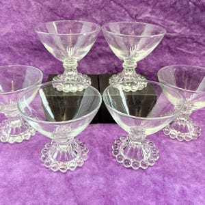May include: Six clear glass dessert or champagne coupe glasses. Each glass has a round bowl, a short stem, and a decorative base with small, round details. The glasses are arranged on a purple surface.